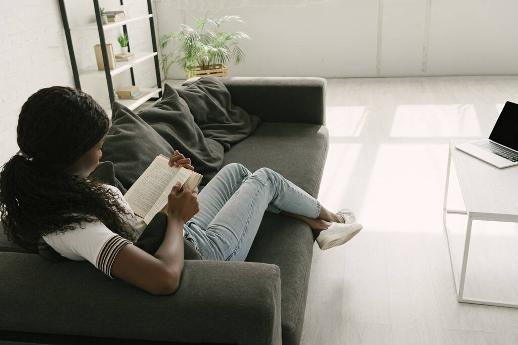 vista de ángulo alto del freelancer afroamericano descansando en el sofá mientras lee el libro cerca de la mesa con el ordenador portátil
 - Foto, Imagen