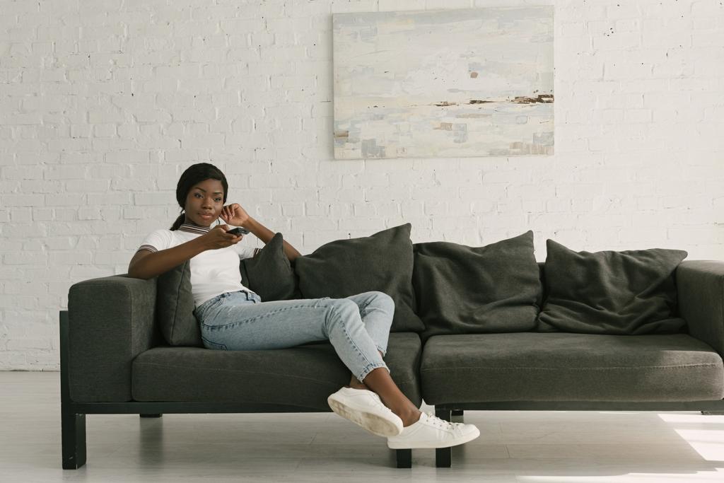 atractivo afroamericano chica sosteniendo tv mando a distancia mientras sentado en sofá en casa
 - Foto, imagen