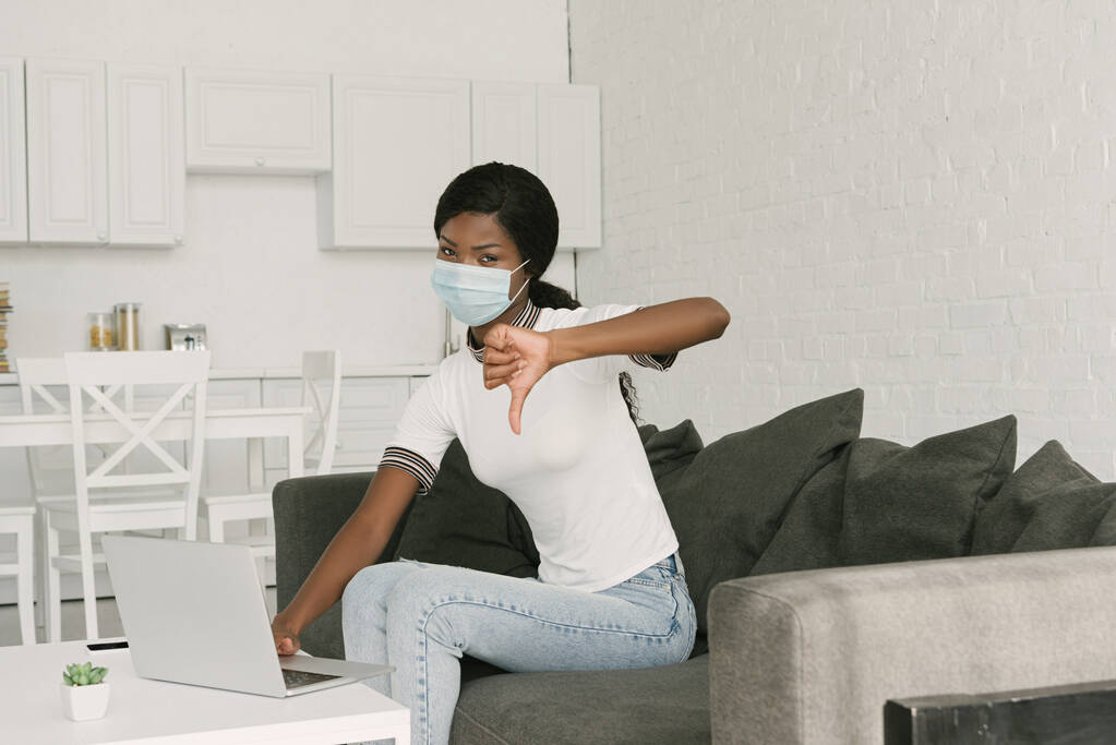 joven afroamericano freelancer en máscara médica mostrando el pulgar hacia abajo mientras está sentado cerca de la computadora portátil en la cocina
 - Foto, imagen