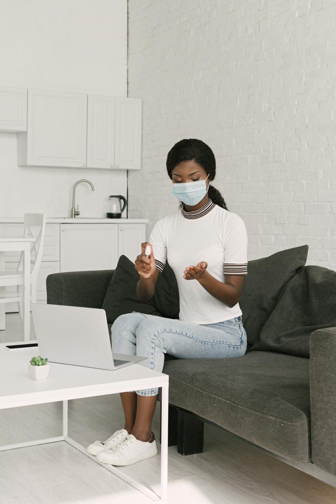Genç Afro-Amerikalı, tıbbi maskeli, mutfaktaki dizüstü bilgisayarın yanında otururken eline antiseptik sıkarak serbest çalışıyor. - Fotoğraf, Görsel