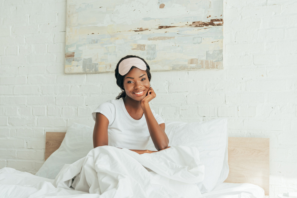 Весела афроамериканська дівчина з маскою для сну сидить у ліжку і посміхається перед камерою. - Фото, зображення
