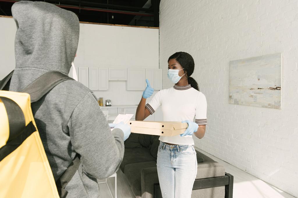 Afrikanerin in medizinischer Maske und Latexhandschuhen nimmt Kisten mit Pizza vom Lieferanten - Foto, Bild