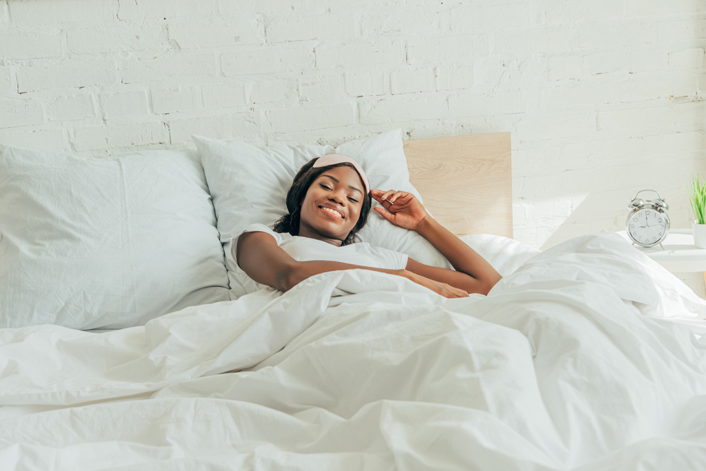 щаслива афроамериканська дівчина зі сном маска на лобі лежить у ліжку і посміхається на камеру
 - Фото, зображення