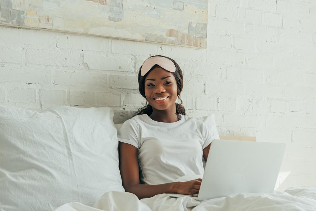 szczęśliwy afrykański freelancer z maską snu na czole za pomocą laptopa w łóżku i uśmiecha się do kamery - Zdjęcie, obraz