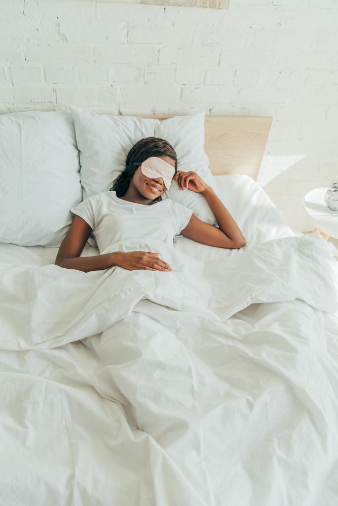 высокий угол обзора улыбающейся африканской девушки, лежащей в постели в маске для сна
 - Фото, изображение