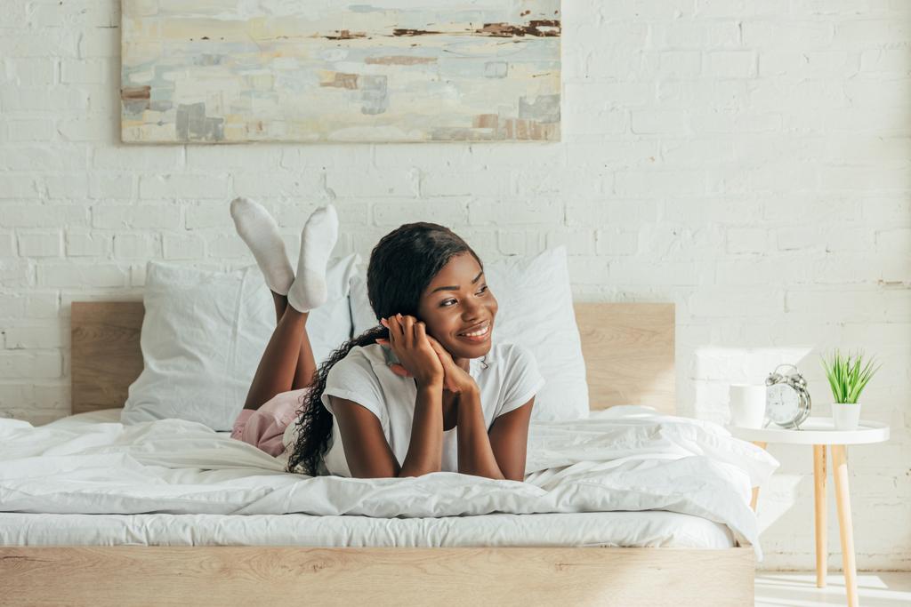 Χαρούμενη Αφρικάνα Αμερικανίδα ξαπλωμένη στο κρεβάτι, χαμογελώντας και κοιτώντας αλλού - Φωτογραφία, εικόνα