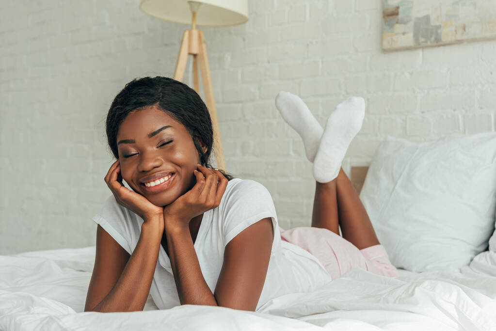 счастливая африканская американская девушка улыбается, расслабляясь в постели с закрытыми глазами
 - Фото, изображение