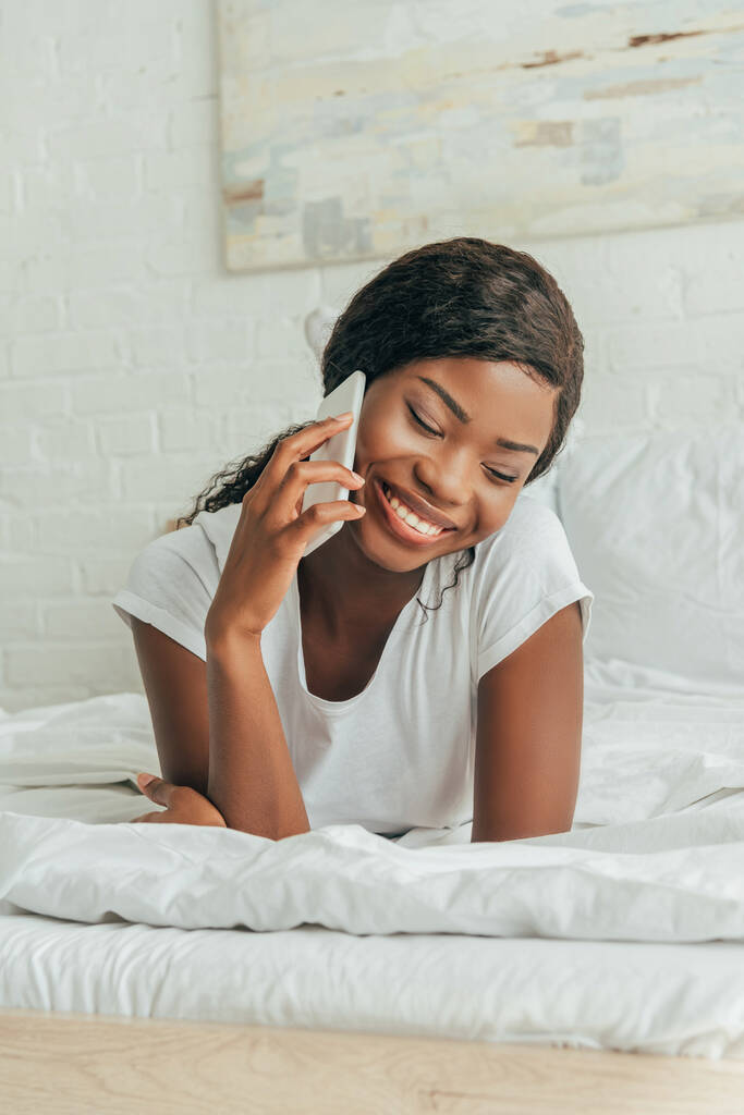 felületi szinten vidám afro-amerikai lány beszél okostelefonon, miközben feküdt a fehér ágynemű - Fotó, kép