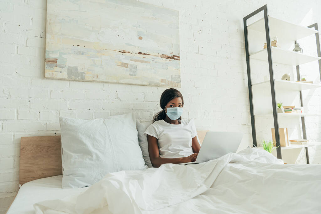 Африканский американский фрилансер в медицинской маске сидит в постели с ноутбуком и смотрит в камеру
 - Фото, изображение