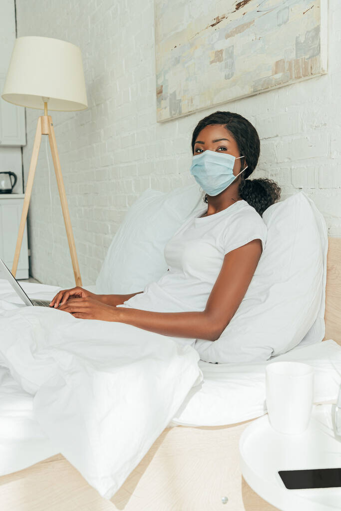Tıbbi maskeli Afrikalı Amerikalı serbest yazar dizüstü bilgisayarla yatakta oturup kameraya bakıyor. - Fotoğraf, Görsel