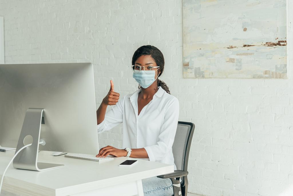 joven afroamericano freelancer en máscara médica mostrando el pulgar hacia arriba mientras está sentado cerca del monitor de la computadora en casa
 - Foto, Imagen