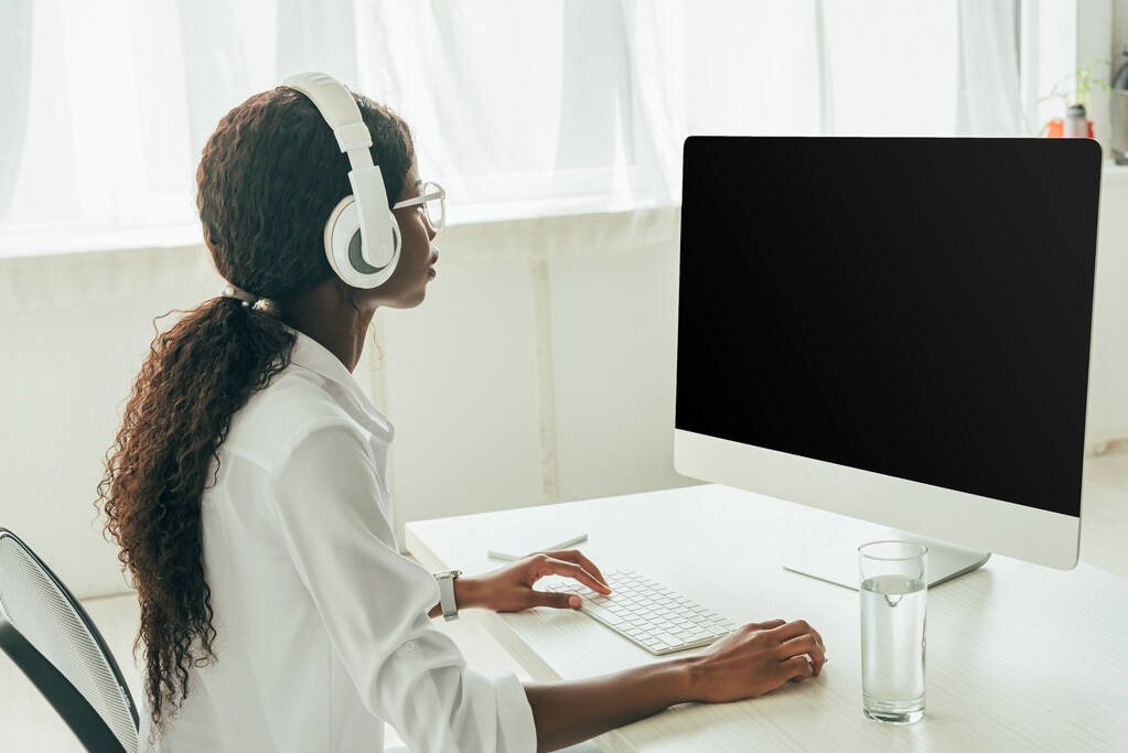 jeune freelance afro-américain dans les écouteurs sans fil en regardant le moniteur d'ordinateur avec écran blanc
 - Photo, image