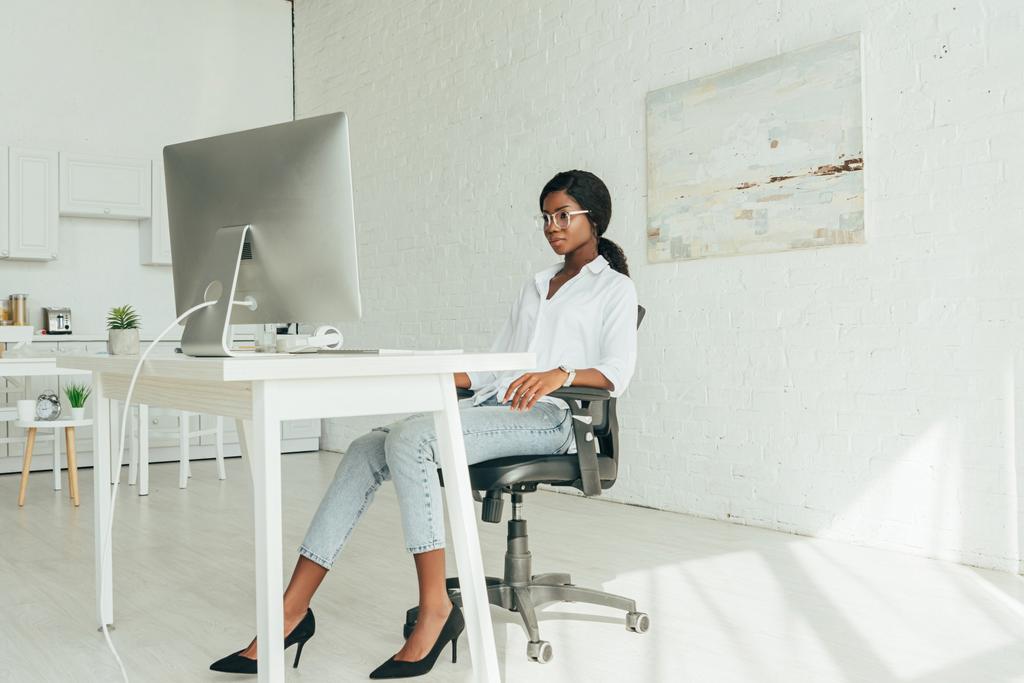 junge afrikanisch-amerikanische Freelancerin in weißer Bluse, Jeans und hochhackigen Schuhen, die zu Hause auf den Computermonitor schauen - Foto, Bild
