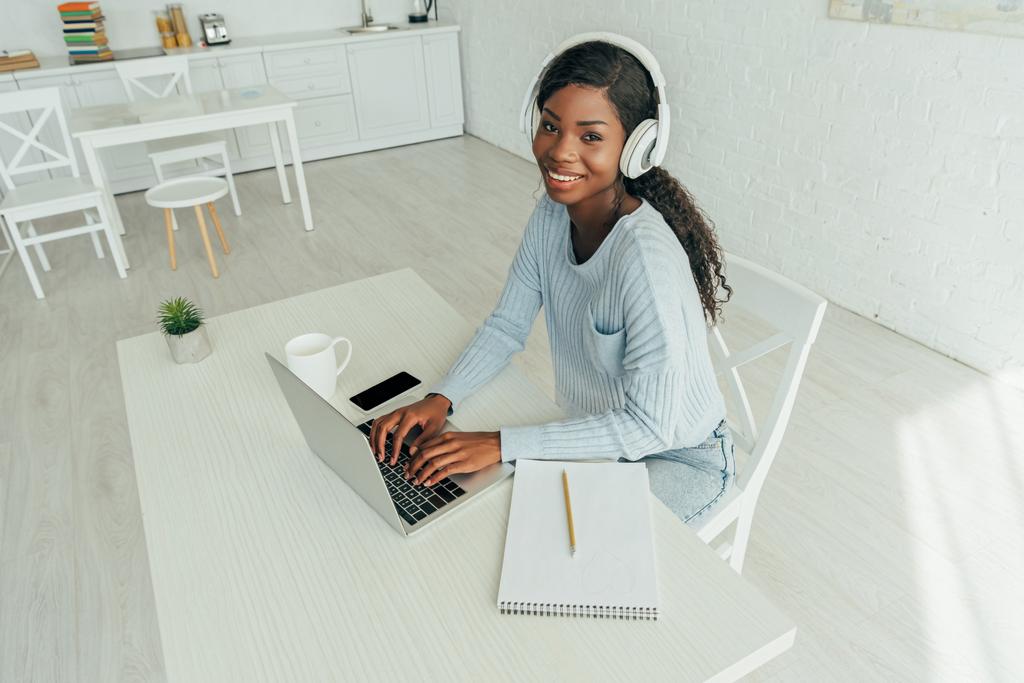 vista ad alto angolo di sorridente afro-americano freelance in cuffie wireless che lavorano su laptop in cucina
 - Foto, immagini