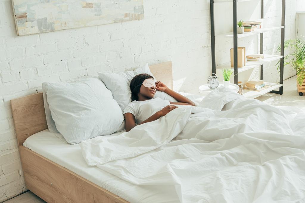 Африканская американка в маске для сна спит на белых кроватях в просторной спальне
 - Фото, изображение