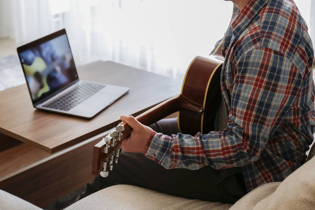 Μουσικός φοιτητής hipster σε καρό καρό πουκάμισο εξάσκηση ακουστική κιθάρα άσκηση, ανάγνωση σημειώσεων από φορητό υπολογιστή. Ο άνθρωπος παρακολουθεί μαθήματα μουσικής στο σπίτι κατά τη διάρκεια της καραντίνας. Φόντο, κοντινό - Φωτογραφία, εικόνα