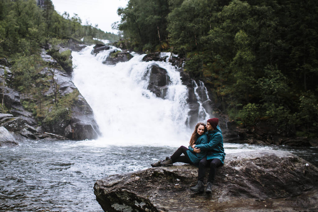 A couple of lovers in green raincoats, sitting on a rock, against the backdrop of a waterfall - Photo, Image