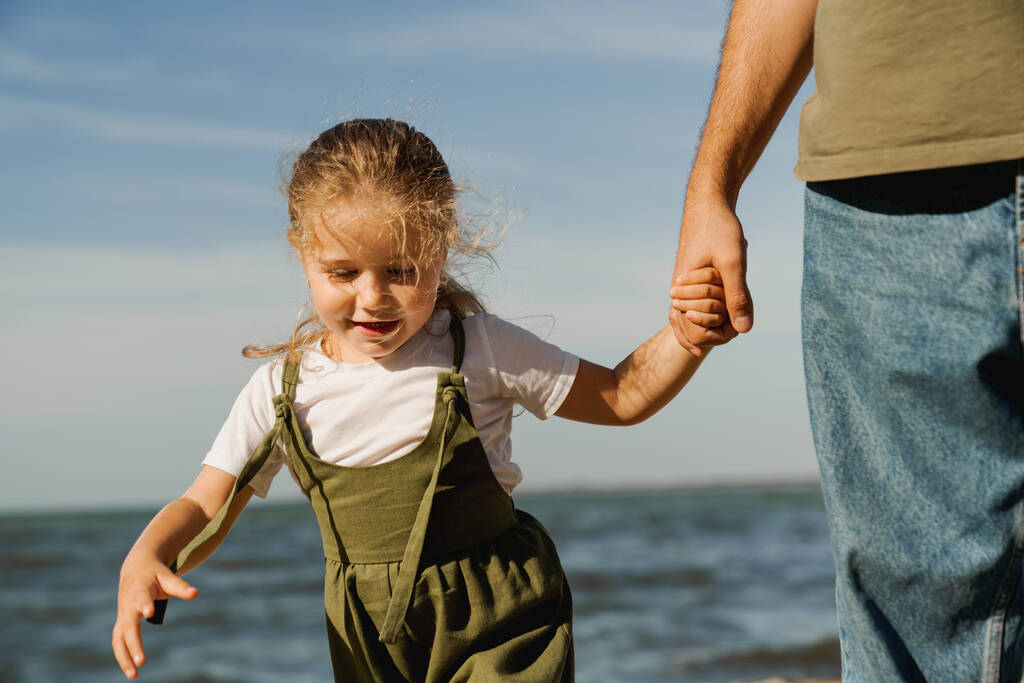 Αξιολάτρευτο κοριτσάκι που κρατάει το χέρι του πατέρα της ενώ περπατάει στην ηλιόλουστη παραλία έξω. - Φωτογραφία, εικόνα