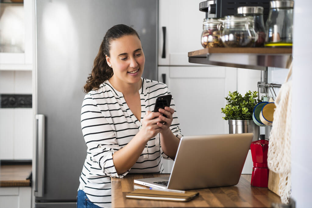 Usmívající se mladá žena pomocí svého smartphonu pracuje s notebookem v kuchyni svého bytu. Práce doma a zůstat doma. - Fotografie, Obrázek