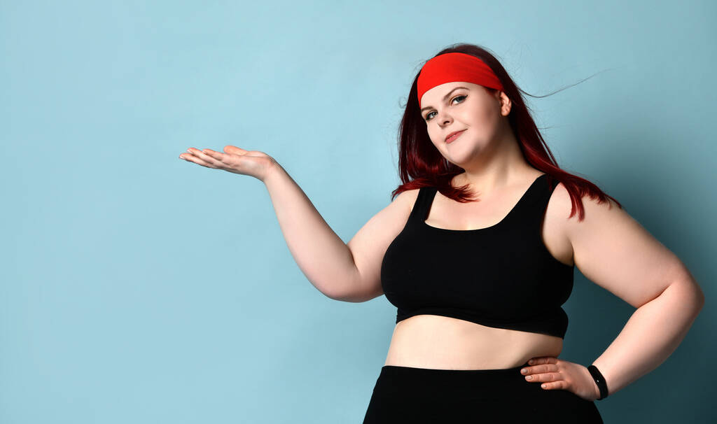 Übergewichtige Rothaarige in rotem Stirnband, schwarzem Oberteil und Leggings. Sie hält etwas auf ihrer Handfläche und posiert auf blauem Hintergrund - Foto, Bild