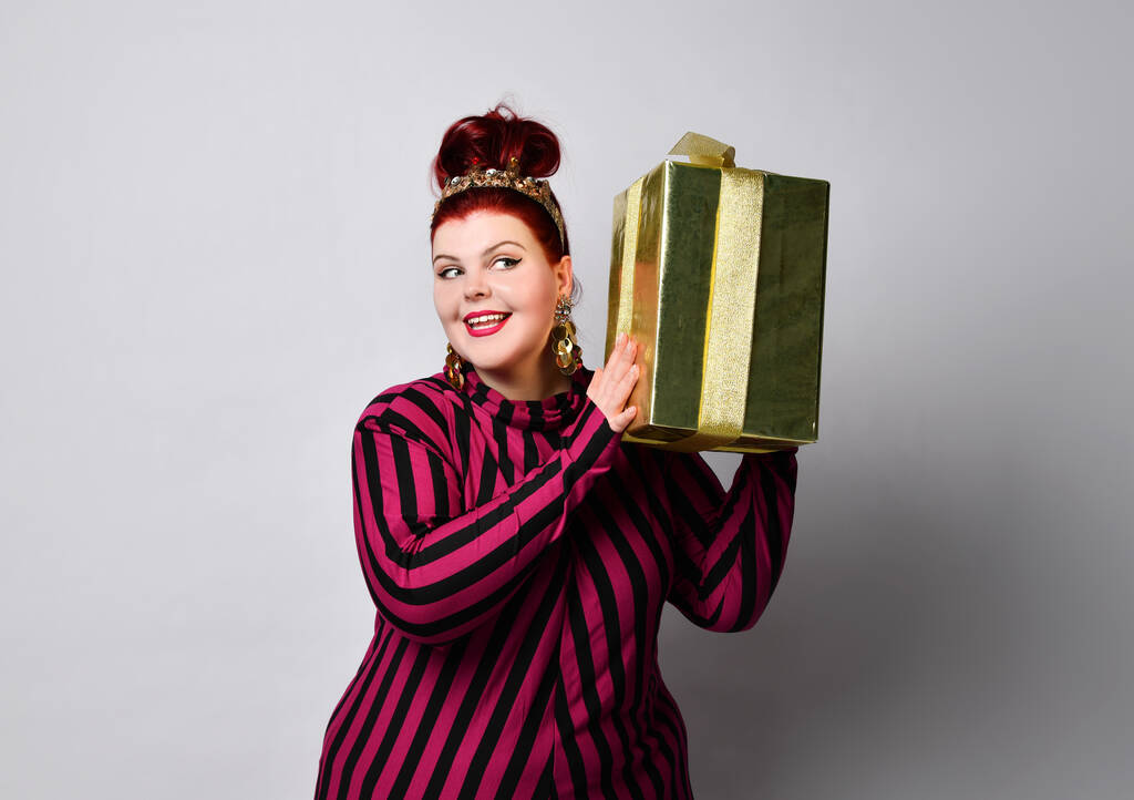 Plus size model in black and purple striped dress, crown and earrings. Holding golden gift box, smiling, posing isolated on white - Photo, Image