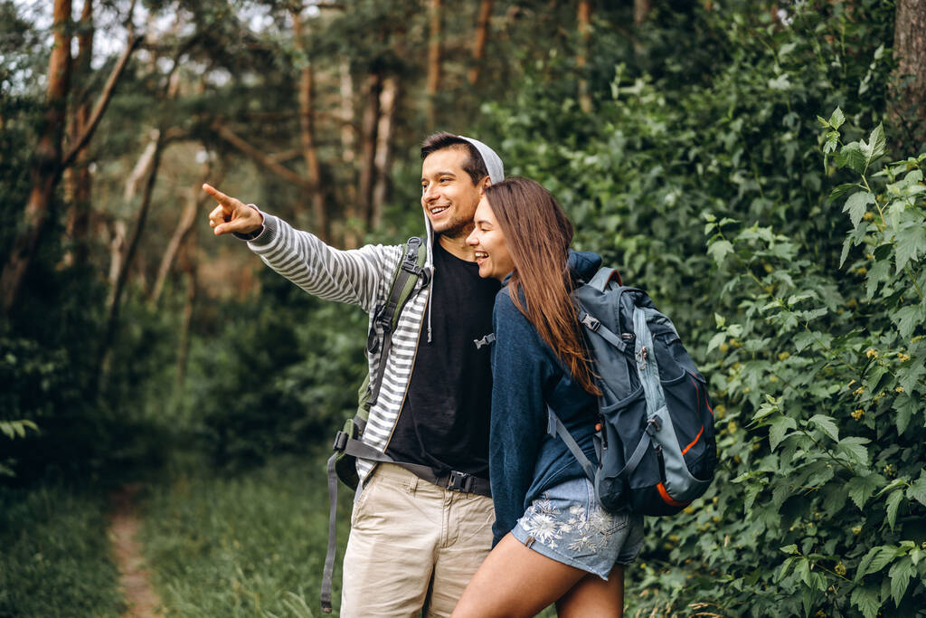Νεαρό ζευγάρι με σακίδια στην πλάτη χαμογελώντας και περπατώντας στο δάσος, απολαύστε τον περίπατο. - Φωτογραφία, εικόνα