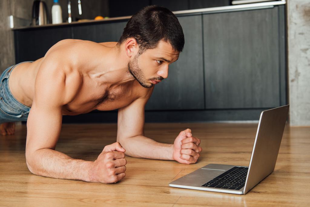 hombre musculoso haciendo ejercicio tablón mientras mira el entrenamiento en línea en el ordenador portátil
  - Foto, Imagen