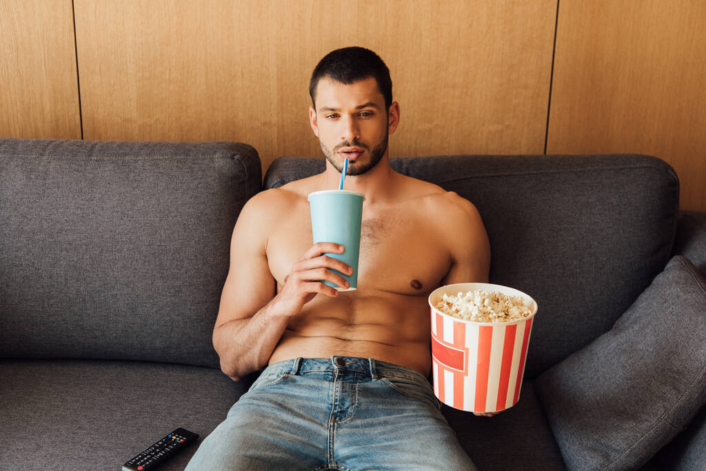 Hemdloser Mann hält Popcorn-Eimer und trinkt Limo neben Fernbedienung auf Sofa - Foto, Bild