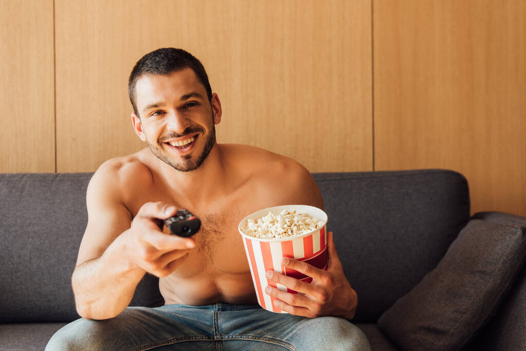 selectieve focus van gelukkige en shirtloze man met afstandsbediening en popcorn emmer  - Foto, afbeelding
