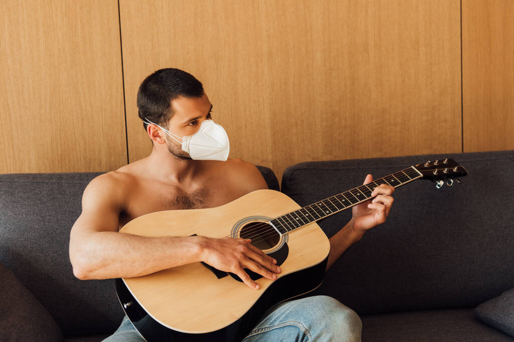 hombre muscular en máscara médica tocando la guitarra acústica en la sala de estar
  - Foto, imagen