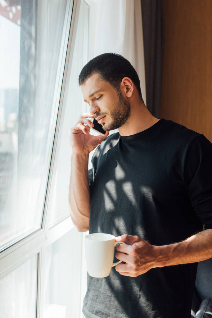 hombre barbudo sosteniendo la taza mientras habla en el teléfono inteligente en casa
 - Foto, imagen