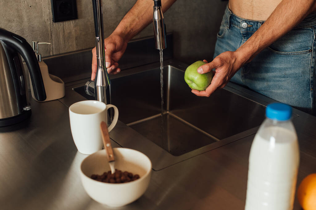 台所に水を注ぐと蛇口の近くにリンゴを持っている男のクロップドビュー  - 写真・画像