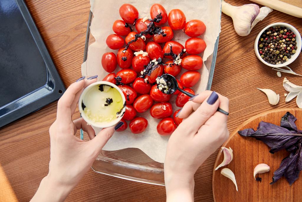 Vista recortada de la mujer preparando tomates cerca de la bandeja del horno y tabla de cortar con ingredientes sobre fondo de madera
 - Foto, Imagen