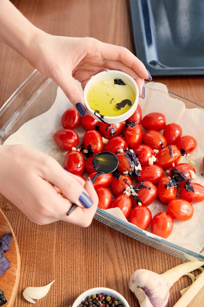 Vista cortada da mulher adicionando azeite de oliva aos tomates com ingredientes perto da bandeja do forno no fundo de madeira
 - Foto, Imagem