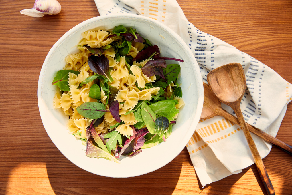 Bovenaanzicht van kom met pasta salade ingrediënten in de buurt van knoflook, servet en spatels op houten achtergrond - Foto, afbeelding