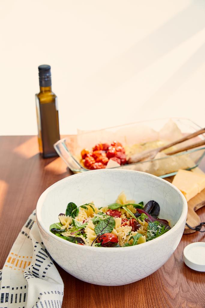 Bol avec salade de pâtes et sel près d'une bouteille d'huile d'olive, plat de cuisson et serviette sur fond bois et beige
 - Photo, image