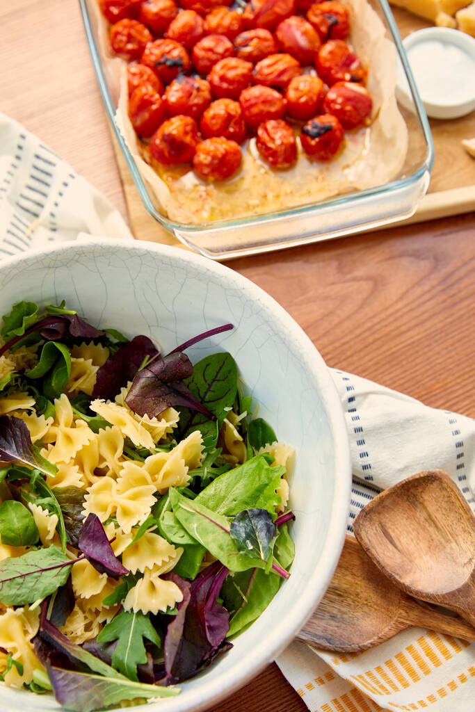 Makarna salatası içerikli kabın yüksek açılı görüntüsü spatula, peçete ve ahşap zemin üzerinde fırın kabında domatesli kesim tahtası. - Fotoğraf, Görsel