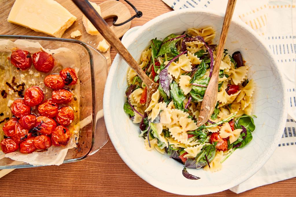 Bovenaanzicht van kom met pasta salade en spatels in de buurt van ingrediënten op snijplank en servet op houten achtergrond - Foto, afbeelding