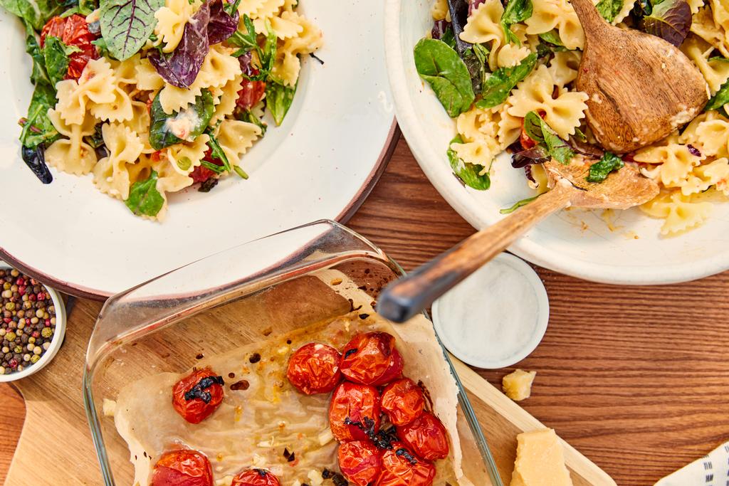 Bovenaanzicht van bord en schaal met pasta salade en spatels in de buurt van snijplank en bakplaat met ingrediënten op houten achtergrond - Foto, afbeelding