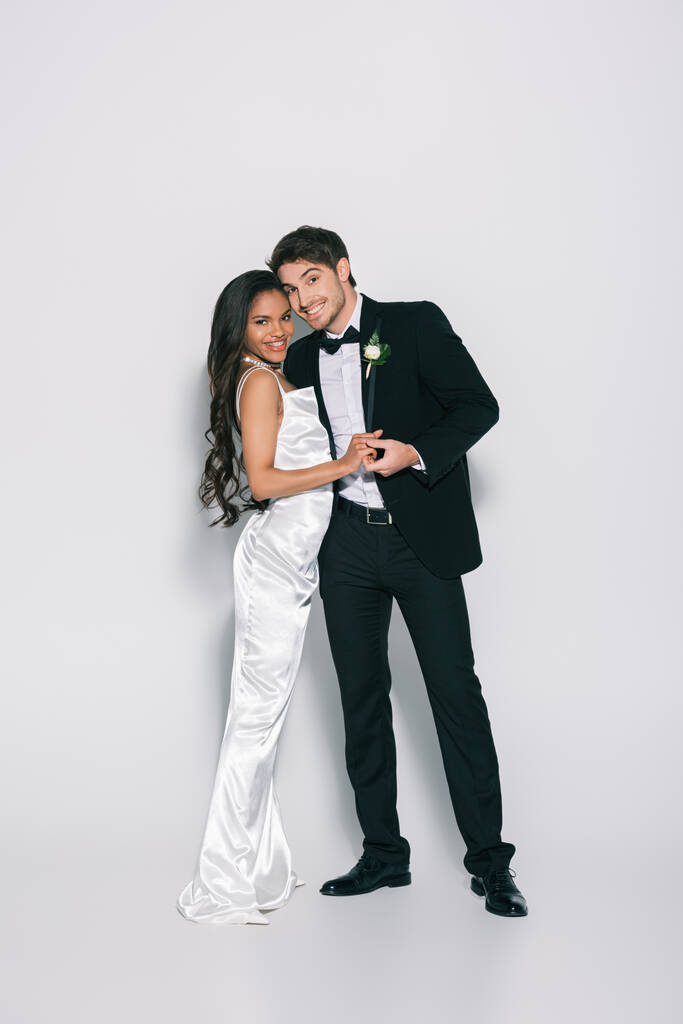 visão de comprimento total de feliz, elegante inter-racial recém-casados de mãos dadas e sorrindo para a câmera no fundo branco
 - Foto, Imagem