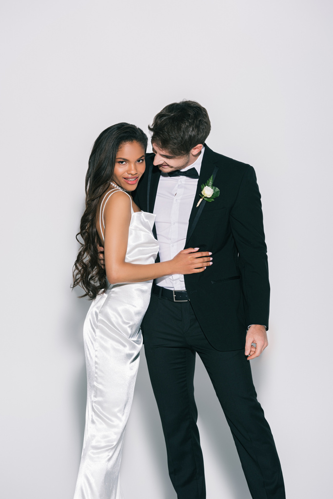 attraktive afrikanisch-amerikanische Braut blickt in die Kamera, während sie eleganten Bräutigam auf weißem Hintergrund berührt - Foto, Bild