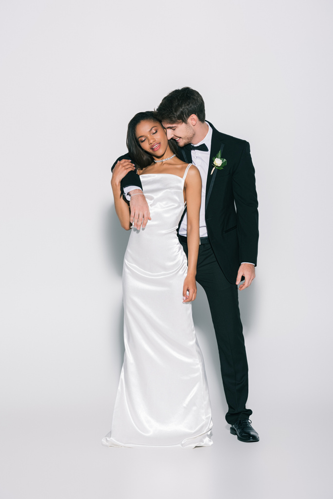 vista completa de feliz, elegante interracial recién casados sonriendo mientras posan sobre fondo blanco
 - Foto, Imagen