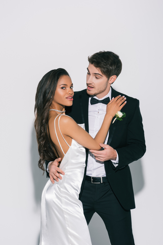 elegante, noiva americana africana atraente olhando para a câmera ao tocar belo noivo no fundo branco
 - Foto, Imagem