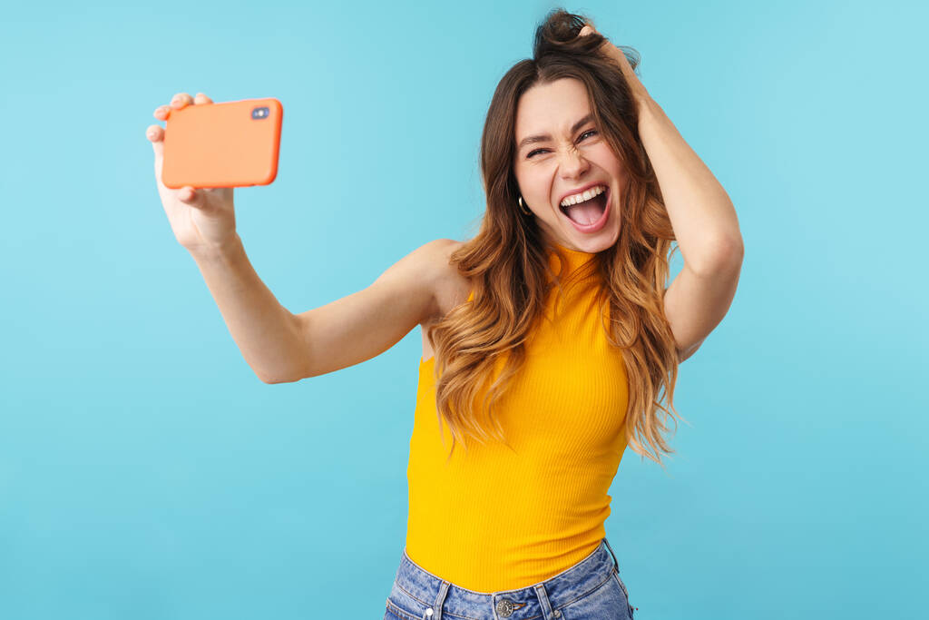 Portrait de belle femme caucasienne joyeuse souriante et prenant selfie photo sur téléphone portable isolé sur fond bleu en studio
 - Photo, image