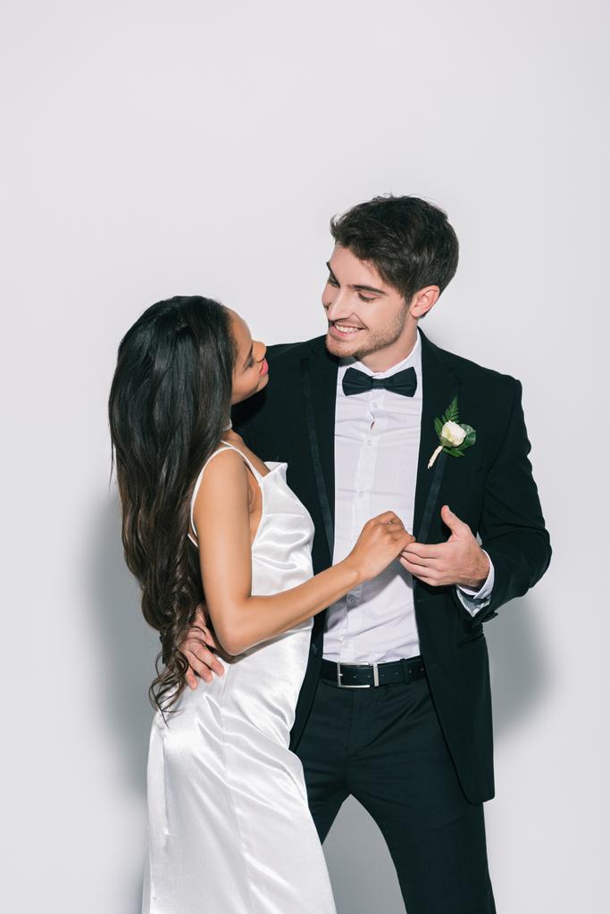 happy, elegant interracial newlyweds looking at each other on white background - Photo, Image