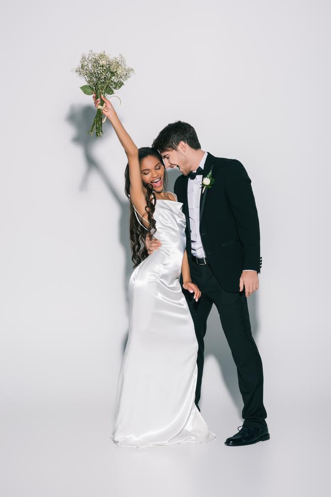 повний вигляд щасливої афроамериканської нареченої, що тримає весільний букет у піднятій руці біля усміхненого нареченого на білому тлі
 - Фото, зображення