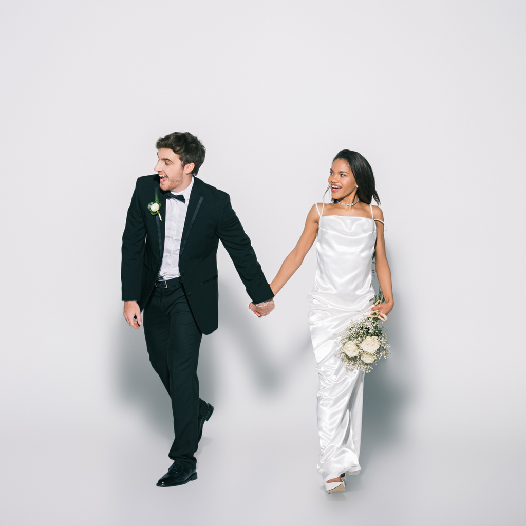 笑顔でエレガントな異人種間の新婚夫婦が白い背景を見て歩きながら手を取り合っています - 写真・画像