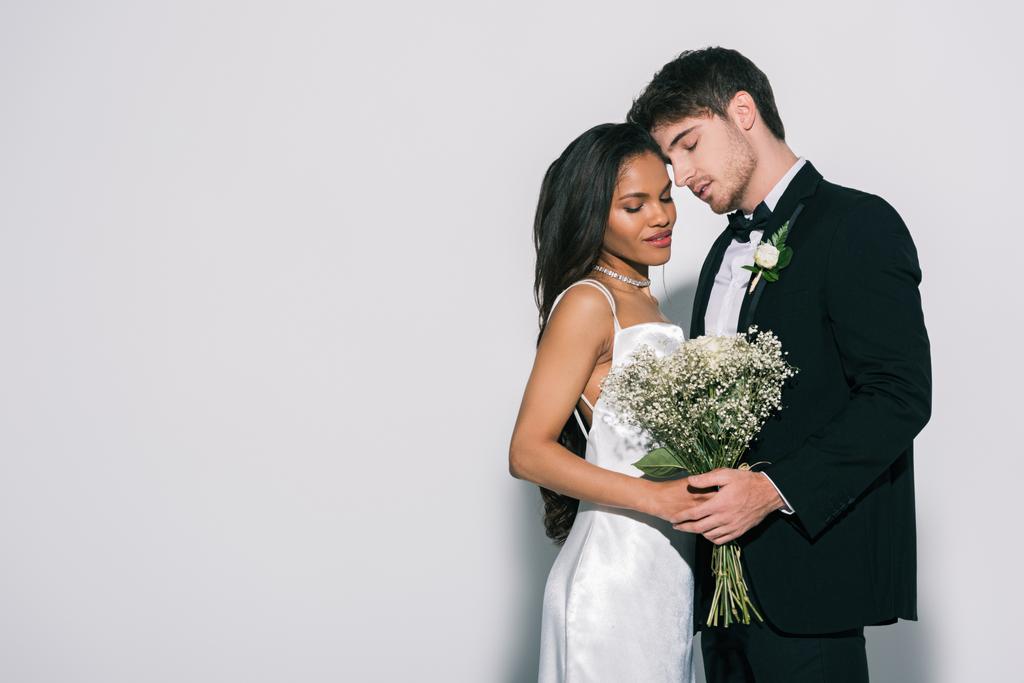 knappe bruidegom en aantrekkelijke Afrikaanse Amerikaanse bruid met bruiloft boeket staan oog in oog met gesloten ogen op witte achtergrond - Foto, afbeelding
