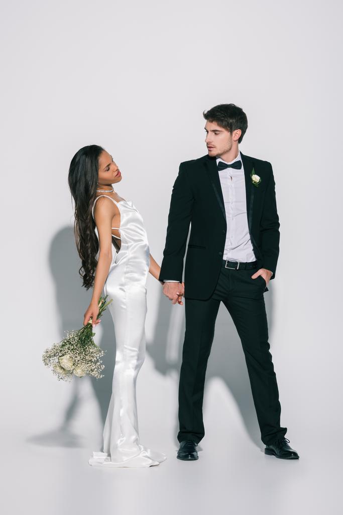 plná délka pohled na elegantní mezirasové novomanželé držící se za ruce a při pohledu na sebe na bílém pozadí - Fotografie, Obrázek