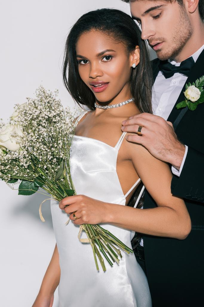 beautiful african american girl with wedding bouquet looking at camera while bridegroom touching her shoulder on white background - Photo, Image
